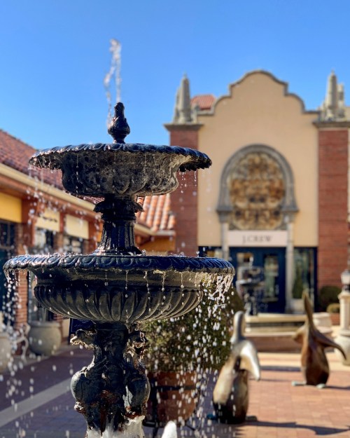 A decorative fountain with water flowing, set in a plaza with shops and sculptures in the background under a clear blue sky.