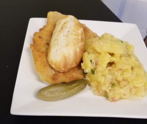 A plate featuring fried fish, a bread roll, creamy potato salad, and a pickle slice on a black table.