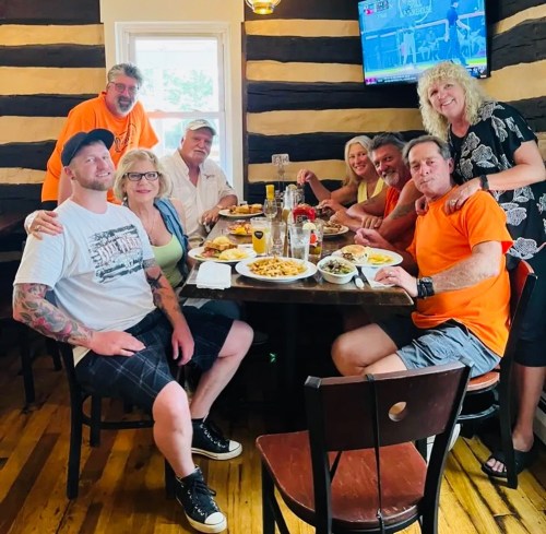 A group of eight people in casual attire gathered around a table with food and drinks, smiling and enjoying their time together.