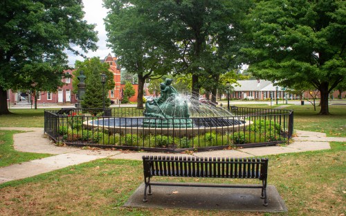 A park scene featuring a fountain with a statue, surrounded by greenery and a bench in the foreground.