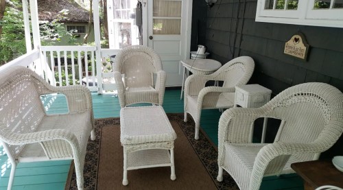A cozy porch with four white wicker chairs and a small table on a teal floor, surrounded by greenery.