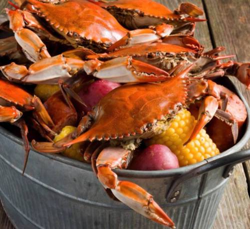 A metal bucket filled with cooked crabs and colorful fruits, including corn and potatoes, on a wooden surface.