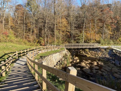 A winding wooden path leads through a forested area with autumn foliage and a small stream.