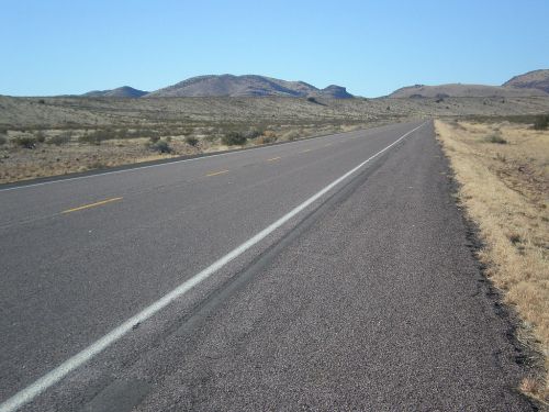 A long, empty road stretches through a desert landscape with distant mountains under a clear blue sky.