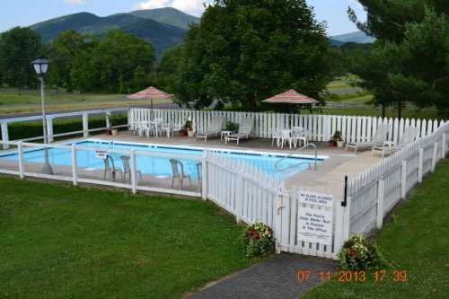 A fenced outdoor pool area with lounge chairs, tables, and umbrellas, surrounded by greenery and mountains in the background.