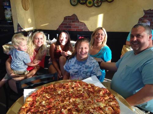 A group of five people, including a child, smiles around a large pizza at a restaurant.