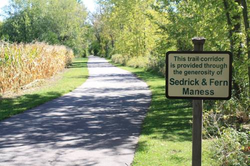 A paved trail surrounded by trees and cornfields, with a sign honoring Sedrick & Fern Maness.