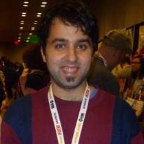 A smiling man with dark hair and a goatee, wearing a striped sweater and a lanyard, stands in a crowded indoor setting.