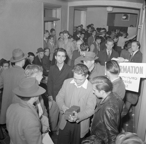 A crowded hallway filled with people, some waiting in line, others talking, with a sign for information visible.