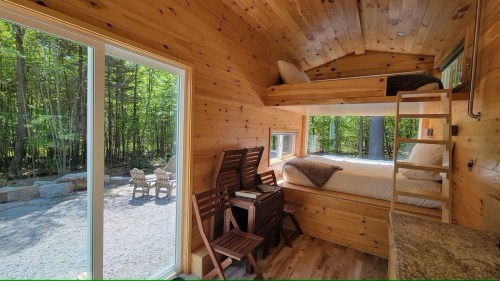 Cozy wooden interior of a tiny cabin with a loft bed, kitchenette, and large windows overlooking a forested area.
