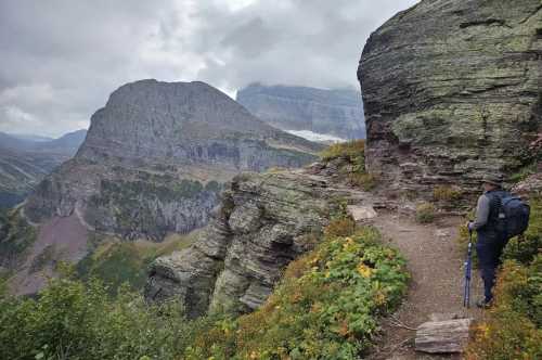 A hiker stands on a rocky trail overlooking a mountainous landscape under a cloudy sky.