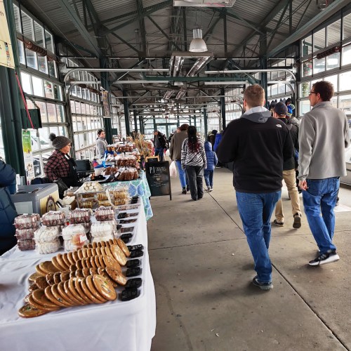 A bustling market scene with people walking past tables of baked goods and snacks in a covered area.