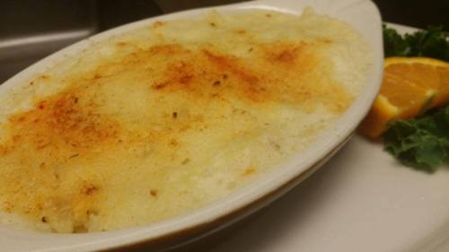 A creamy, baked dish with a golden top, served on a white plate alongside a slice of orange and leafy greens.