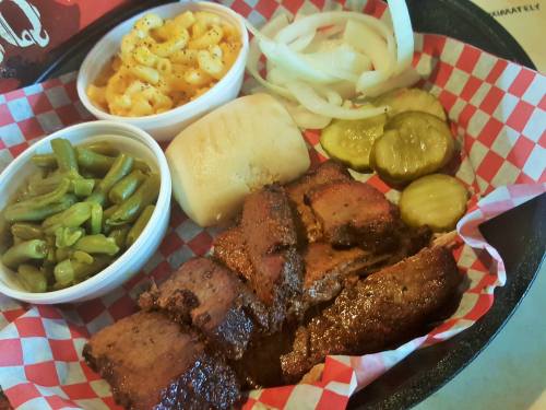 A plate of barbecue brisket with sides of macaroni and cheese, green beans, pickles, onions, and a roll in a basket.