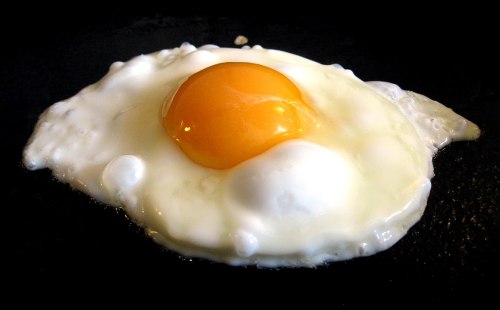 A fried egg with a bright yellow yolk and white albumen, sizzling in a black skillet.
