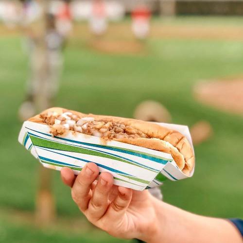 A hand holding a hot dog topped with onions, served in a striped paper wrapper, with a blurred outdoor background.