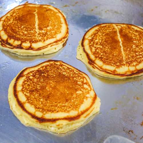 Three golden-brown pancakes cooking on a griddle, with a slightly crispy edge and fluffy texture.