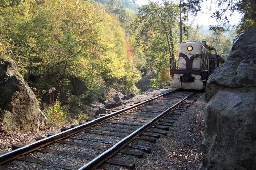 A train approaches on a track surrounded by colorful autumn foliage and rocky terrain.