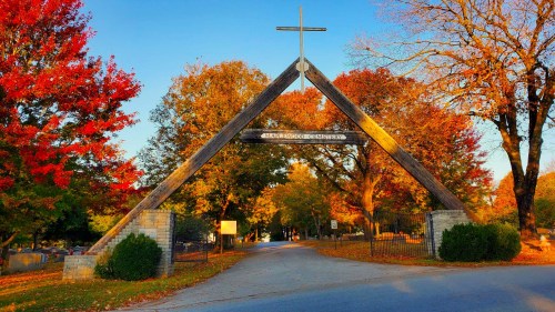A wooden archway with a cross, surrounded by vibrant autumn trees, marks the entrance to a cemetery.