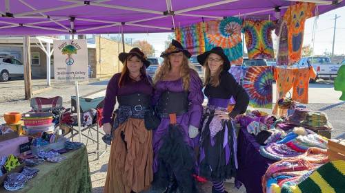 Three women in witch costumes stand at a colorful market stall with various handmade items under a purple canopy.