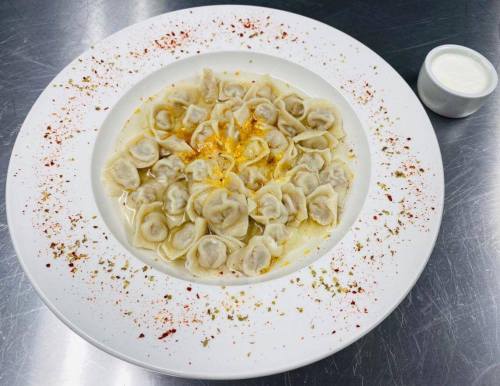 A plate of dumplings in broth, garnished with spices, served with a small cup of sauce on the side.