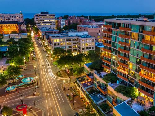 Aerial view of a city street at night, with illuminated buildings, trees, and light trails from moving vehicles.