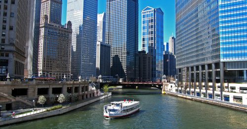 A boat glides along the Chicago River, surrounded by tall skyscrapers under a clear blue sky.