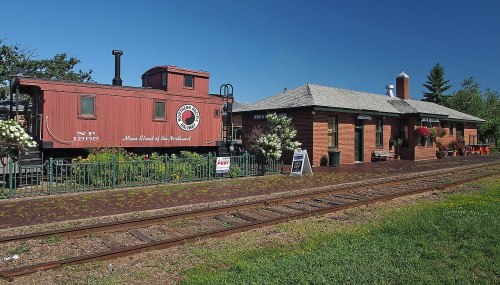 A vintage red caboose next to a brick train station, surrounded by greenery and flowers, with railroad tracks in the foreground.