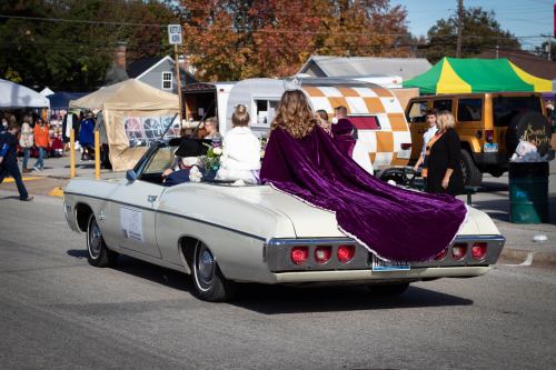 A convertible car with a woman in a purple cape and a girl, driving through a festive outdoor event.