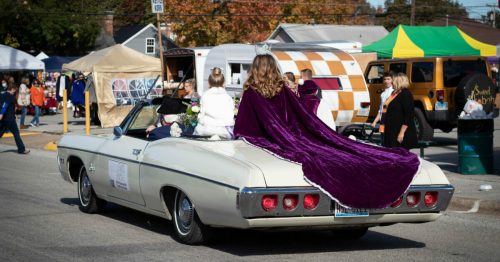 A vintage convertible car with a woman in a purple cape and a girl, driving through a festive outdoor event.