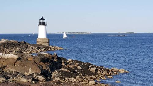 A lighthouse stands on rocky shore, with sailboats in the distance and calm blue waters under a clear sky.