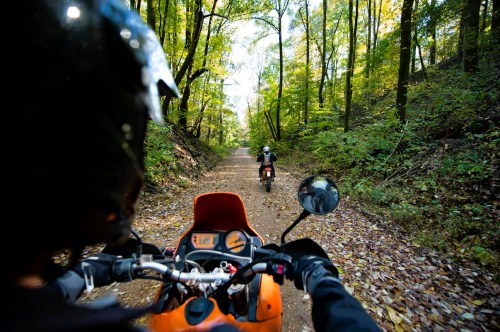 Two motorcyclists ride on a dirt path through a lush, green forest in autumn.