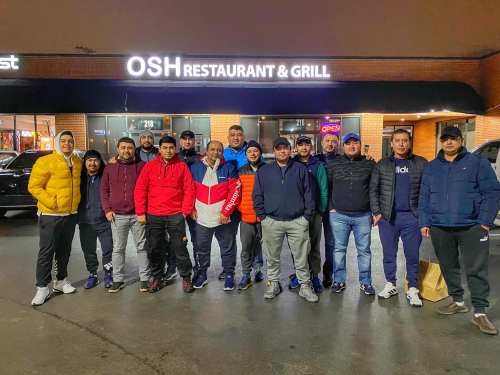 A group of diverse individuals poses outside OSH Restaurant & Grill at night, smiling and dressed in casual winter attire.