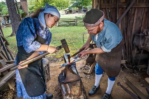 A woman and a man work together at a forge, shaping metal with a hammer and anvil in a rustic outdoor setting.