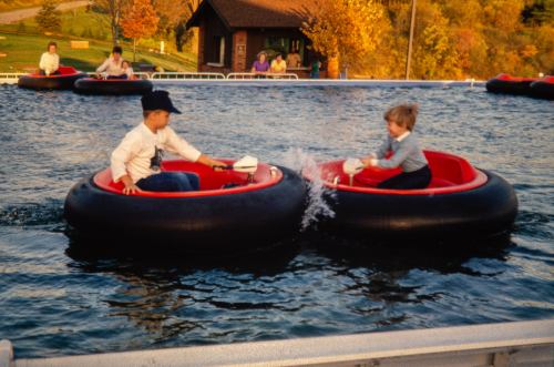 Two children in bumper boats play in a water attraction, splashing each other with water, surrounded by autumn trees.