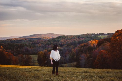 A person in a white poncho stands in a field, overlooking colorful autumn hills under a cloudy sky.