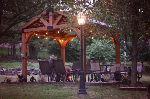 A cozy outdoor gazebo with string lights, surrounded by chairs and a lamp, set in a green garden.