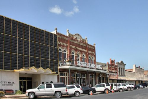 Historic buildings line a street, featuring Sengelmann Hall and City Hall, with parked cars in front under a clear sky.