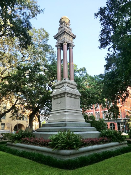 A tall monument with four columns, topped by a globe, surrounded by greenery and flowers in a park setting.