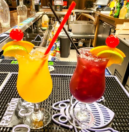 Two colorful cocktails on a bar counter: one orange with a cherry and lime, the other red with an orange slice and cherry.