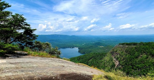 A panoramic view of a lush green landscape with a lake surrounded by mountains under a blue sky with fluffy clouds.