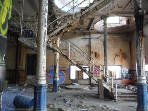 Abandoned interior with a worn staircase, peeling walls, and graffiti, surrounded by debris and a neglected atmosphere.
