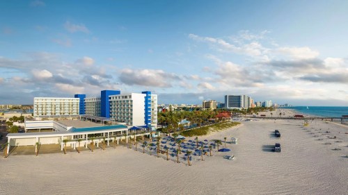 Aerial view of a beach resort with blue buildings, sandy beach, umbrellas, and the ocean in the background.