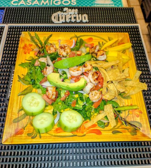 A colorful salad with shrimp, avocado, cucumbers, tomatoes, and tortilla strips on a wooden plate.