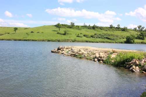 A serene lake with a rocky shore, surrounded by green hills and a blue sky with fluffy clouds.