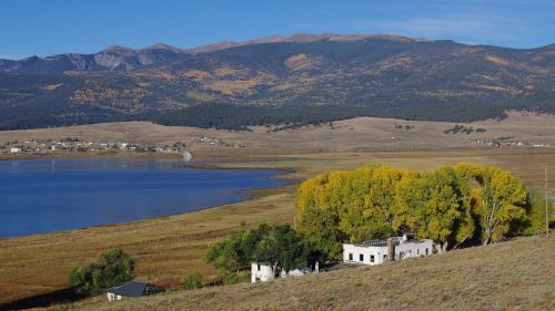A serene landscape featuring a lake, mountains, and a small white building surrounded by autumn-colored trees.