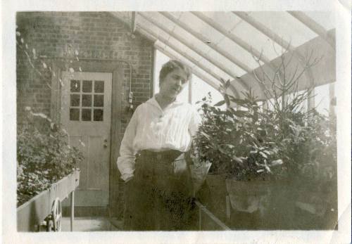 A person stands in a greenhouse, surrounded by potted plants, with a door and window in the background.