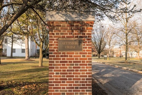 Brick entrance sign for Gettysburg College, surrounded by trees and a pathway on a clear day.