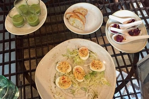 A plate of deviled eggs with greens, accompanied by bread, green shots, and small bowls of dessert on a wooden table.
