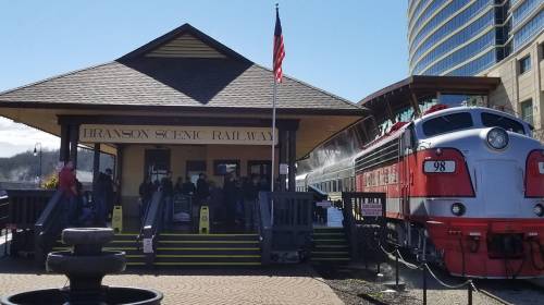 A vintage train at the Branson Scenic Railway station, with passengers boarding and an American flag waving.